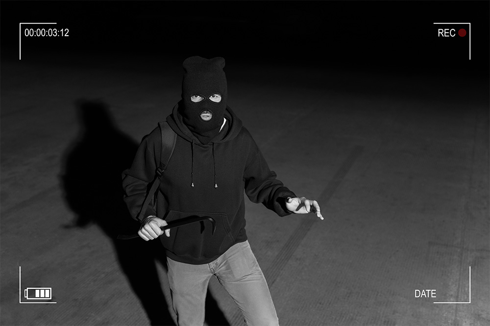 A black and white CCTV screenshot of a man wearing a black hoodie and black ski mask